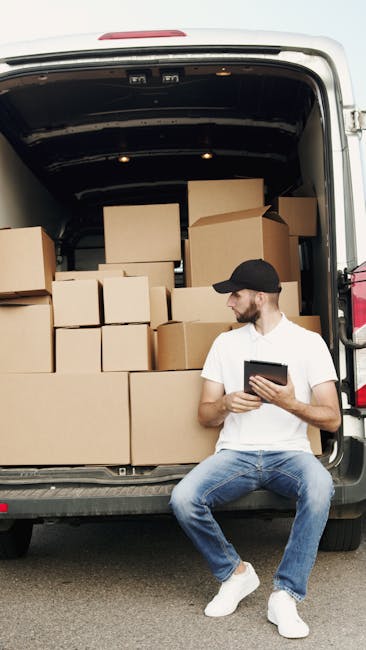 A man wearing a white t-shirt, blue jeans, white sneakers, and a black cap is seated on the pavement beside the open rear doors of a white van, which is loaded with stacked cardboard boxes of various