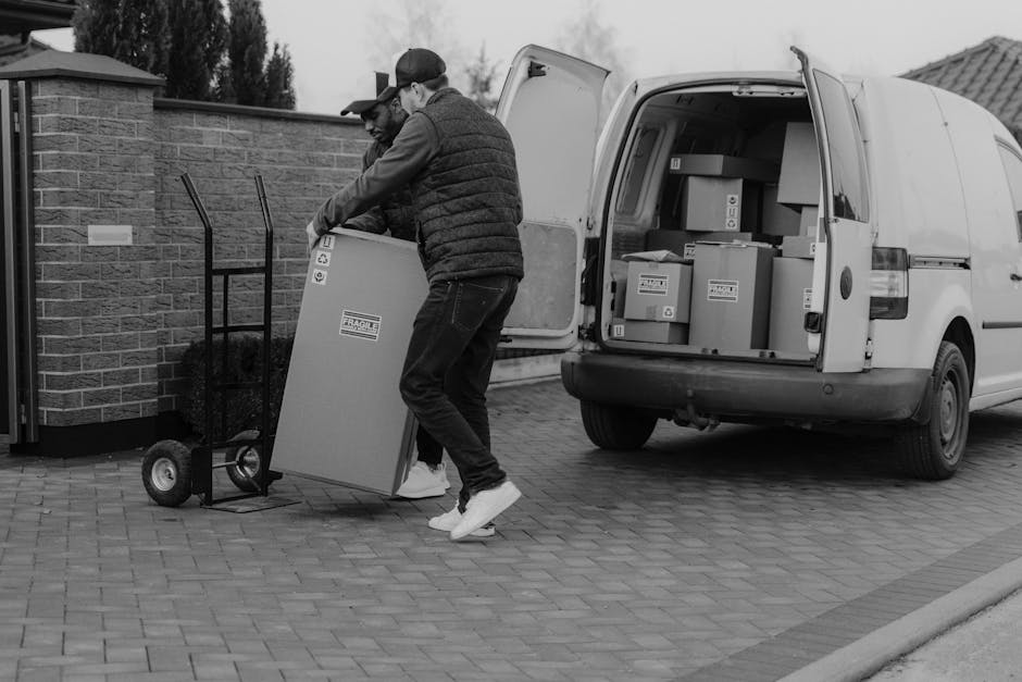 A man wearing a white t-shirt, blue jeans, white sneakers, and a black cap is seated on the pavement beside the open rear doors of a white van, which is loaded with stacked cardboard boxes of various