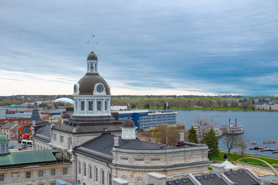 A high-angle view of the rooftop and clock tower of a historic building with a large, round, clock face and a domed turret, set against a partly cloudy sky. The scene captures the building's detailed